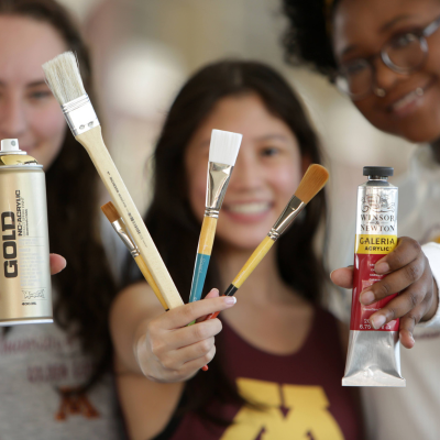 three female students holding up art supplies